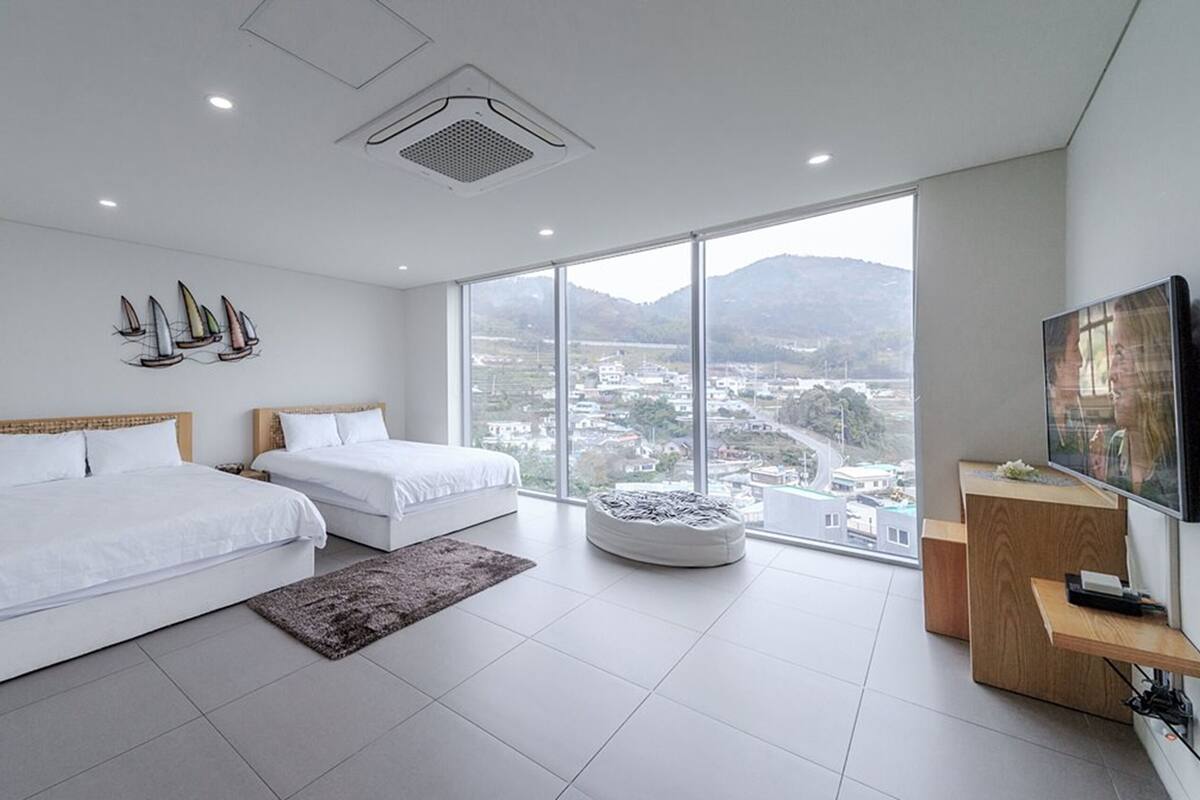 A modern hotel room with two beds, a large window with a city view, a flat-screen TV, and a small wooden side table.