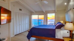 Ein Schlafzimmer mit Bett, Deckenventilator, Fernseher und Blick auf den Sonnenuntergang über dem Meer.