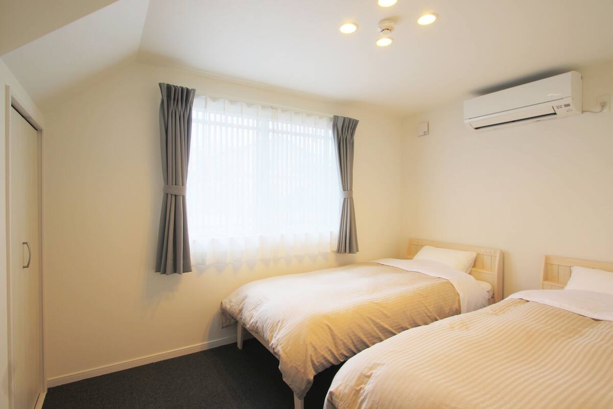 A hotel room with two beds, a window with curtains, an air conditioning unit, and a door.