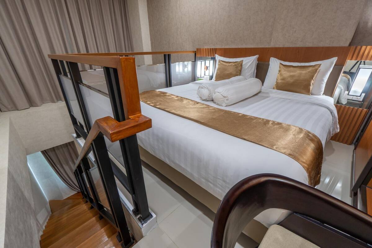 A hotel room with a large bed, a wooden headboard, a gold accent on the bed, a wooden chair, and a staircase.