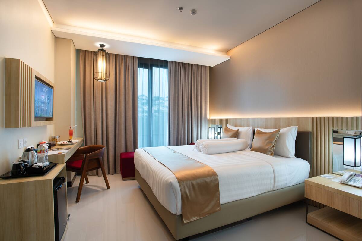 A modern hotel room with a large bed, a desk with a computer, a chair, and a small table with a phone.
