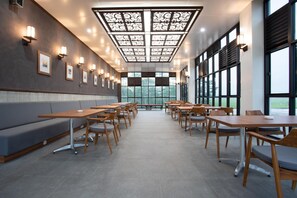 A spacious room with a long bench along one wall, multiple tables and chairs arranged for dining, and large windows providing a view of the outdoors.
