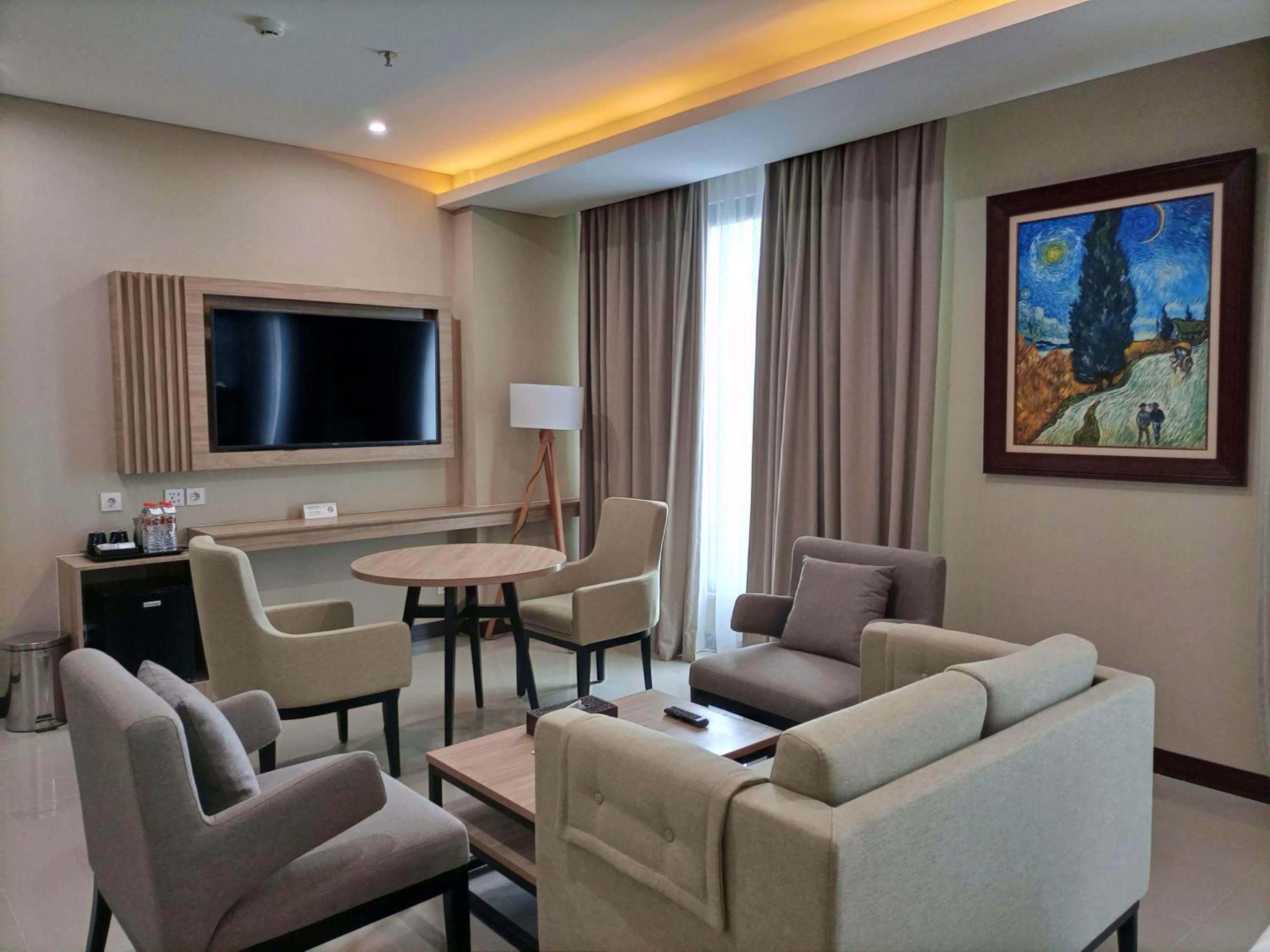A hotel room with a flat-screen TV, a round table, two sofas, a lamp, and a painting on the wall.