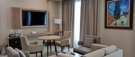 A hotel room with a flat-screen TV, a round table, two sofas, a lamp, and a painting on the wall.