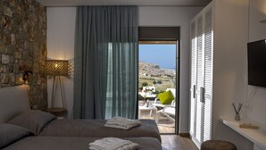 Ein modernes Hotelzimmer mit Steinwand, einem Bett mit zwei Kissen, einer Nachttischlampe, einem Balkon mit Blick auf das Meer und Gebäude sowie einem Fernseher an der Wand.