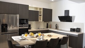 Eine moderne Küche mit einem Edelstahlkühlschrank, einer Mikrowelle und einer Kaffeemaschine. Ein Esstisch mit Stühlen lädt zum Verweilen ein, und ein reichhaltiges Frühstücksbuffet mit Bananen, Saft und Gebäck steht bereit.
