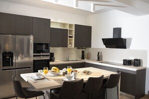 A modern kitchen with a stainless steel refrigerator, a microwave, and a coffee maker. There is a dining table with chairs, and a breakfast spread including bananas, juice, and pastries.