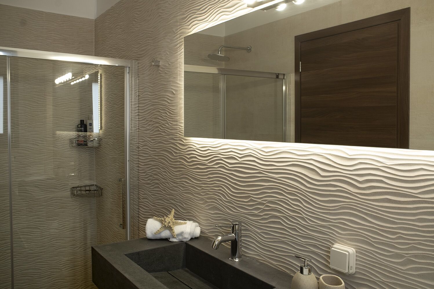 Une salle de bain moderne avec un mur texturé, une douche en verre, un lavabo foncé et un grand miroir.