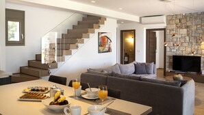 Ein modernes Wohnzimmer mit einem grauen Sofa, einem Esstisch mit Frühstücksutensilien, einer Treppe und einer Steinwand mit Pendelleuchten.