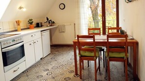 Una cocina pequeña con estufa, horno y una mesa de comedor con sillas. Se ve un ventanal con vistas a árboles y un reloj en la pared.