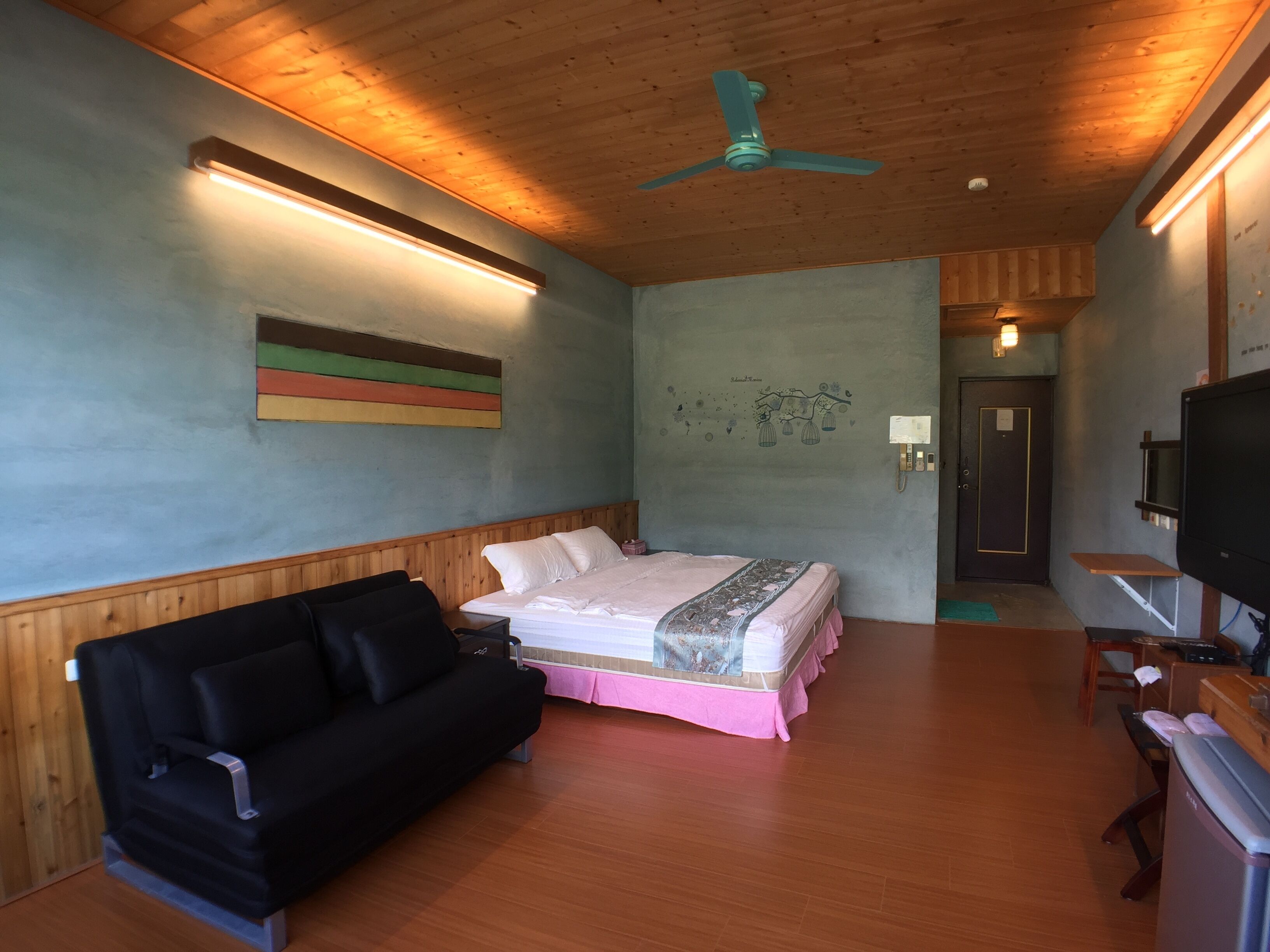 A bedroom with a bed, a black sofa, a ceiling fan, a TV, and a wooden floor.