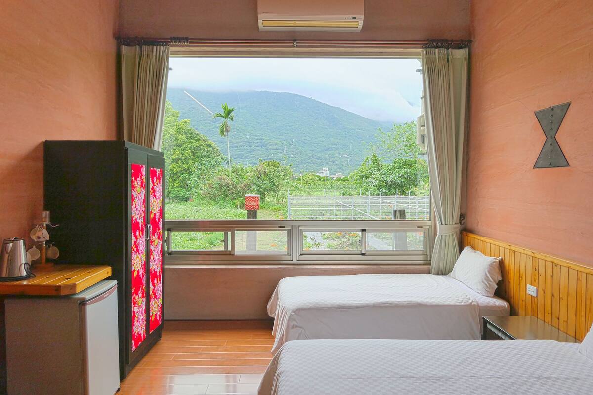 A hotel room with two beds, a small kitchenette, and a window with a view of a mountain and greenery.