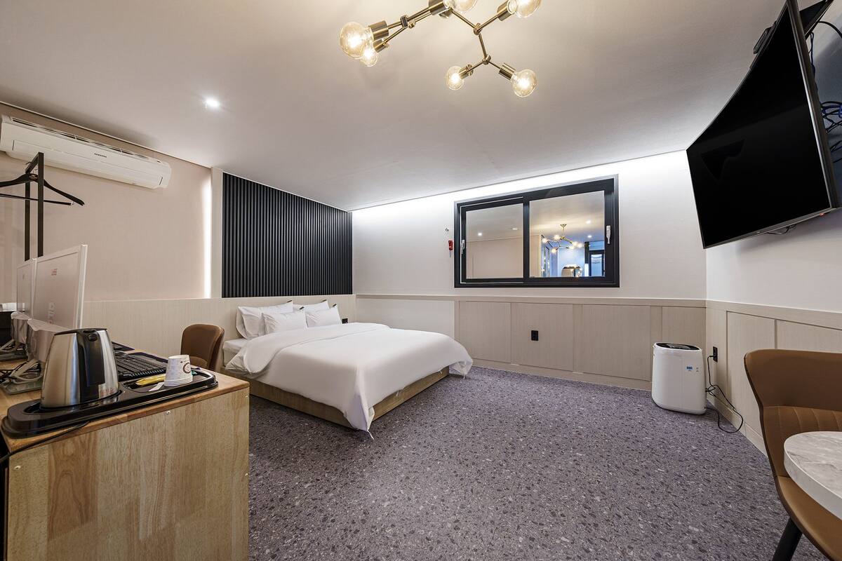 A hotel room with a bed, a desk with a kettle, a TV, and a window.