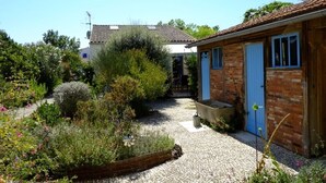 A courtyard with a cobblestone path, a brick building with a blue door, and a garden with various plants and flowers. - Henriette, between beach and market (La Brée-les-Bains)