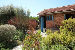 Une maison de campagne en briques, avec un toit en tuiles, une porte bleue et un jardin où poussent diverses plantes et arbustes.