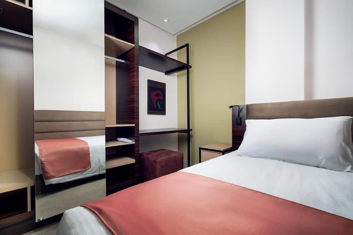 A hotel room with a bed, a wardrobe, a small table, and a wall-mounted shelf.