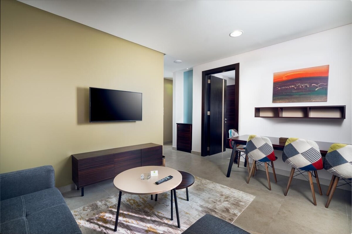 A modern living room with a flat-screen TV, a sofa, a dining table with chairs, and a painting on the wall.