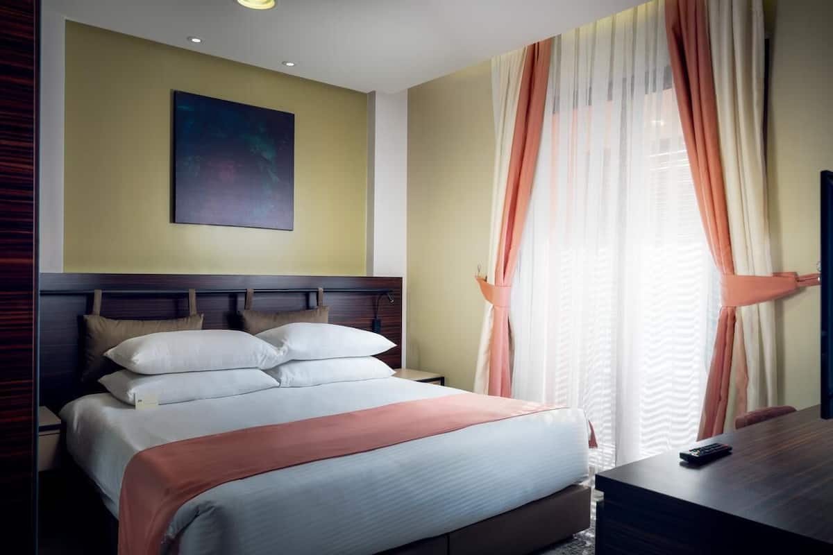 A hotel room with a bed, a nightstand, a desk, a chair, a painting on the wall, and a window with curtains.