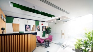 A modern reception area with a wooden reception desk, two pink chairs, a small table with a plant, and a large window providing natural light. - AFAM Residence (Dammam)