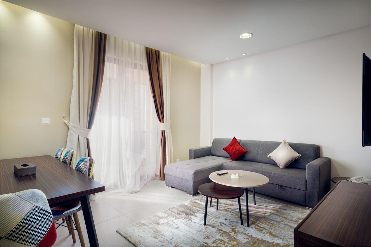 A modern living room with a sofa, a coffee table, a dining table with chairs, and a large window with sheer curtains.
