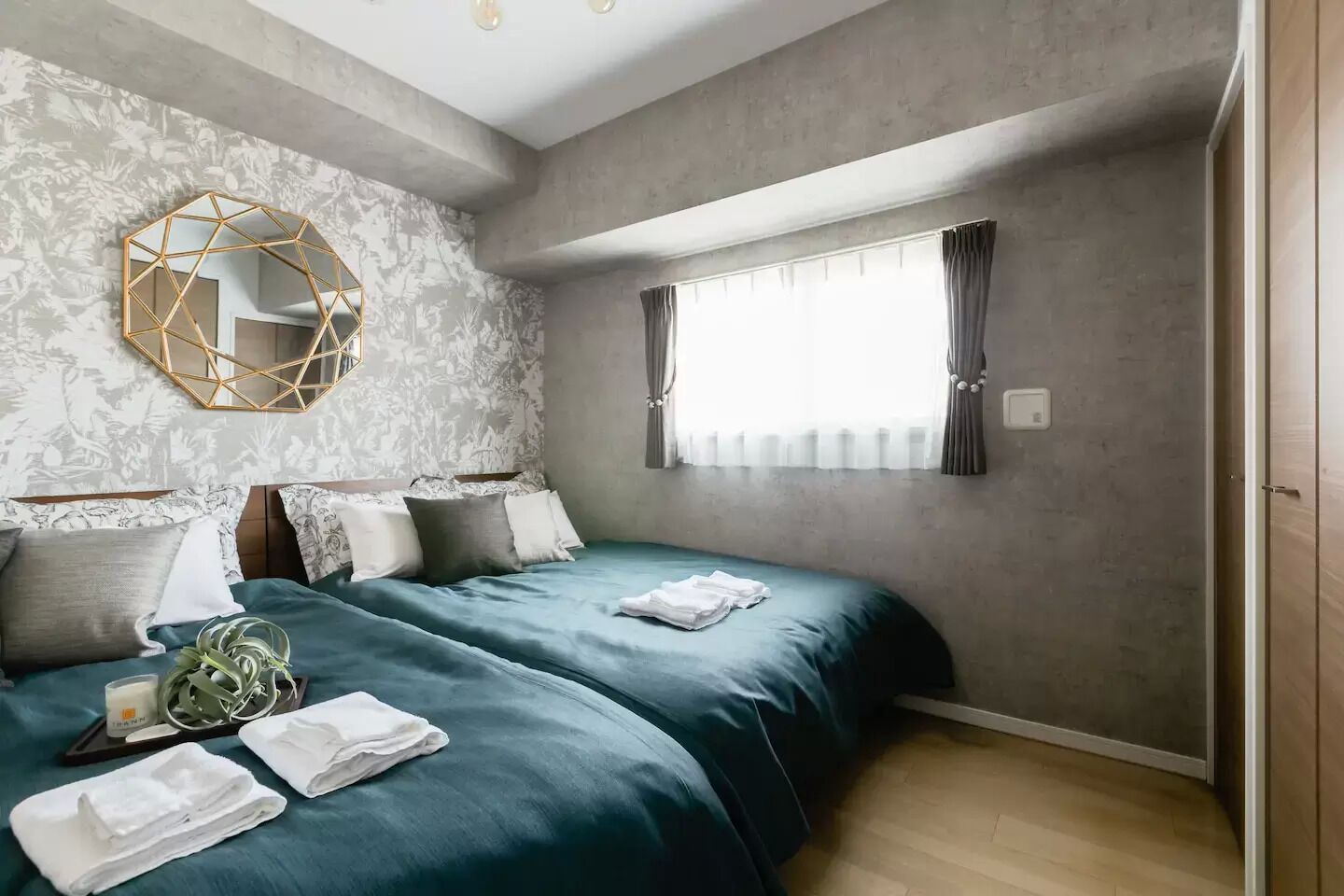 A bedroom with two beds, a geometric mirror, a window with curtains, and a patterned wallpaper.