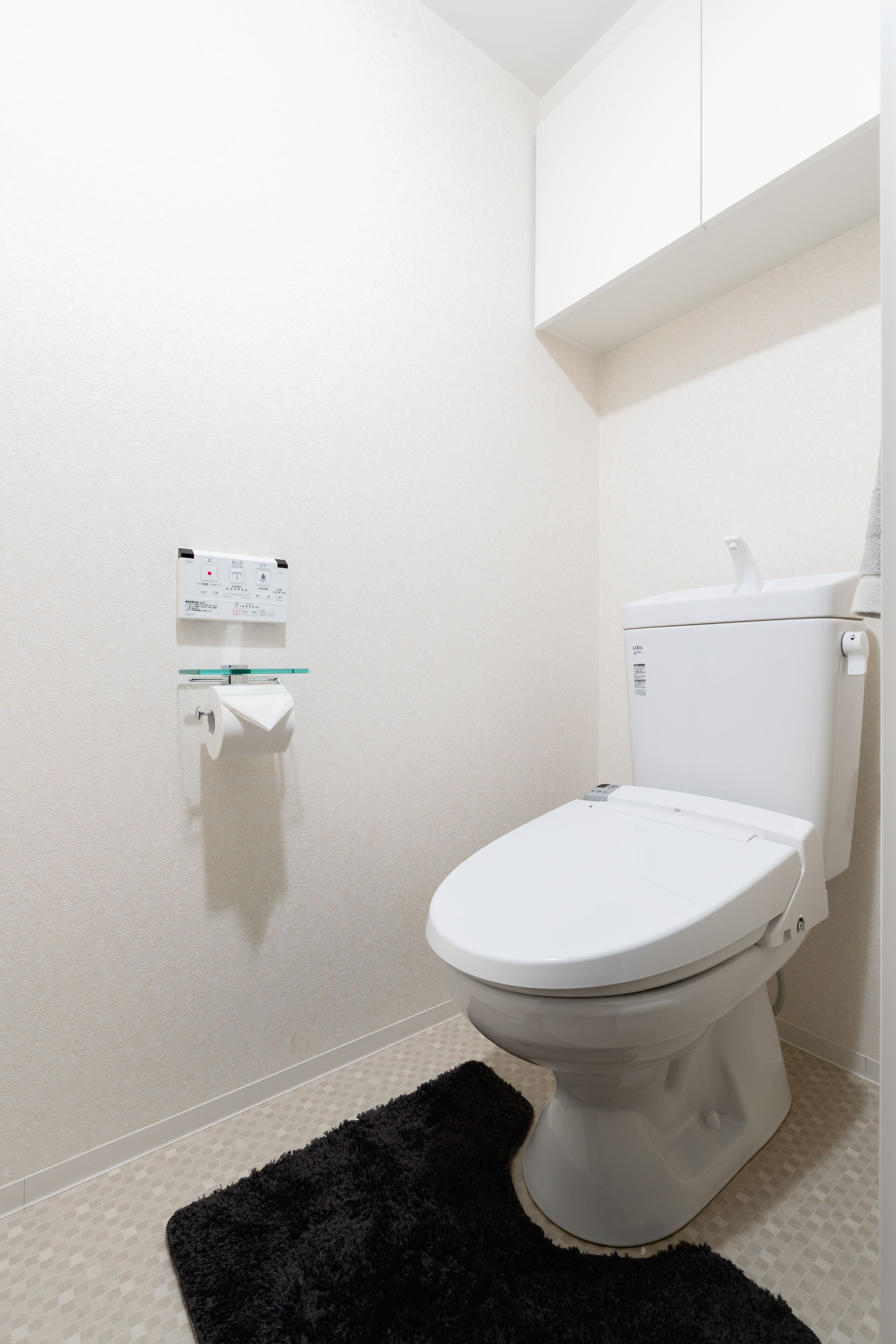 Een kleine, schone badkamer met een wit toilet, een toiletrolhouder met glazen deurtjes en een zwart vloerkleed.