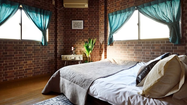 Een kamer met een bakstenen muur, een bed met een wit en grijs dekbed, twee ramen met teal gordijnen, een klein bureau met een lamp en een plant.