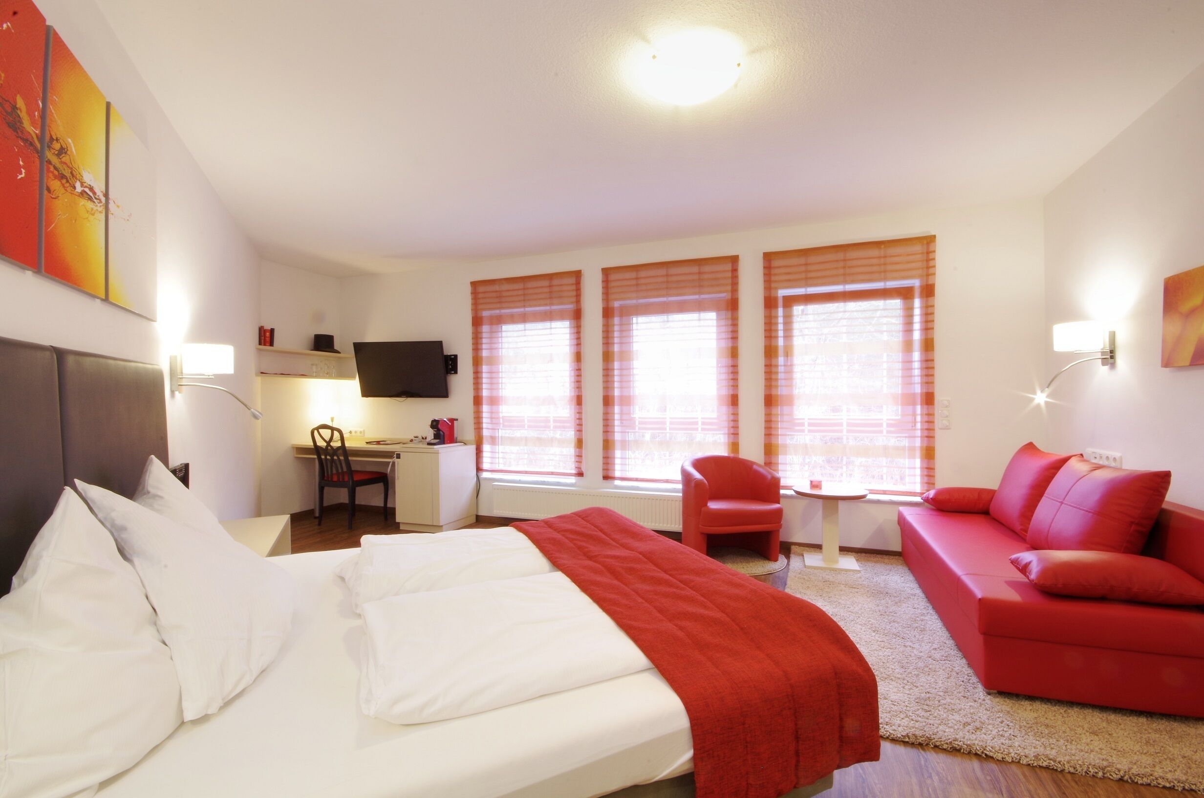 A hotel room with a bed, a red sofa, a desk with a chair, and a TV.
