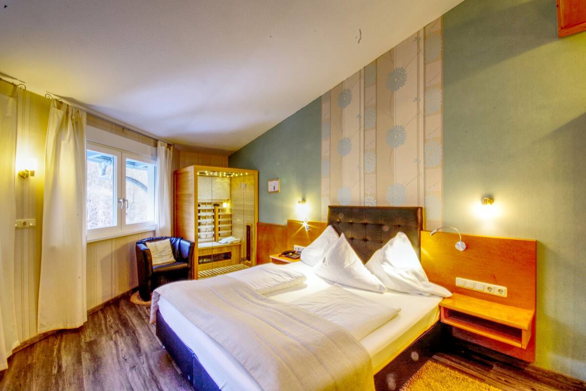 Quarto de hotel com uma cama grande, cabeceira de madeira, mesa de cabeceira, luminária e uma janela com cortinas.