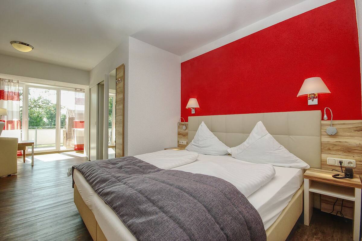 Quarto de hotel com uma cama, mesa de cabeceira, parede de destaque vermelha e vista através de uma porta de vidro.