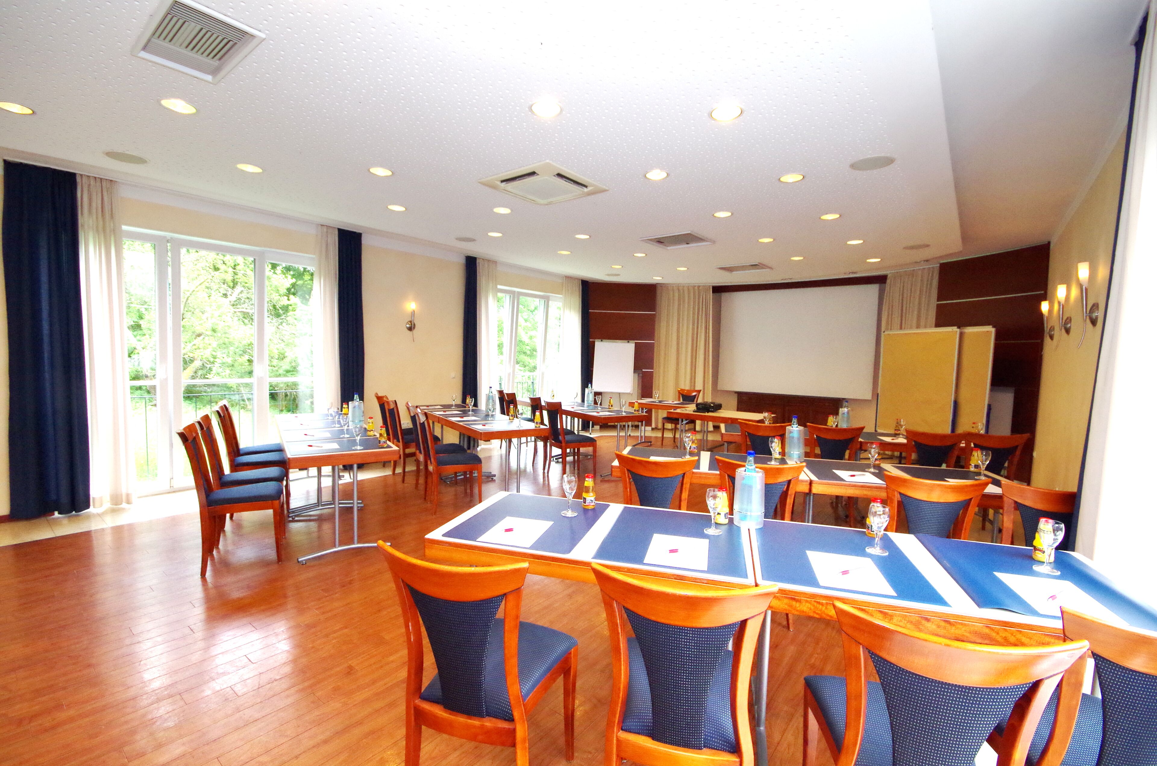 A conference room with multiple tables and chairs, large windows, and a projector screen.