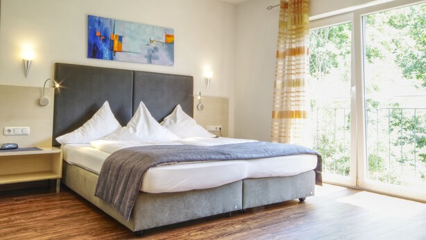 A modern bedroom with a large bed, a nightstand, a wall-mounted lamp, and a painting. - Hotel Jakob (Gross-Umstadt)