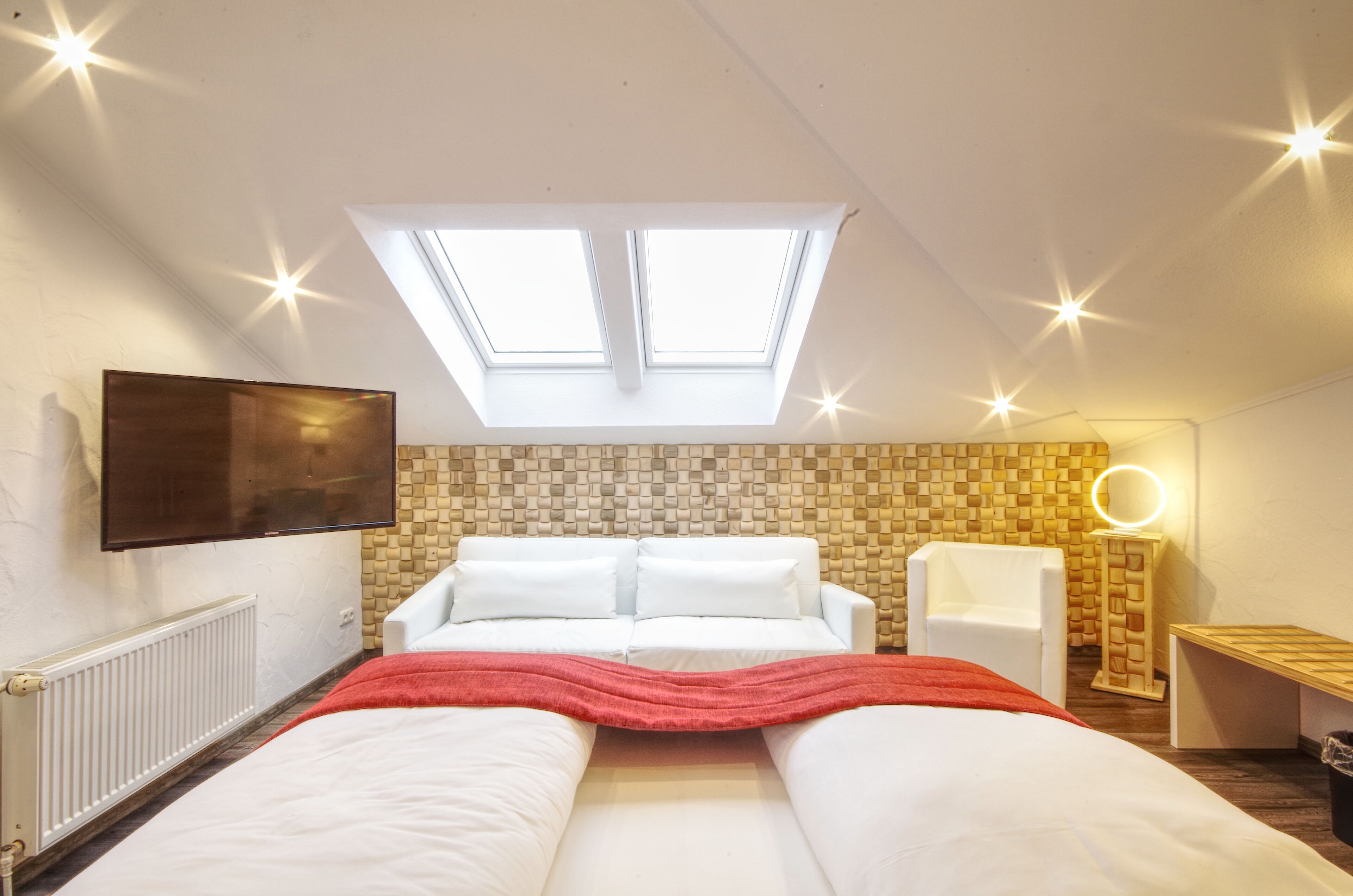 A bedroom with a skylight, a bed, a TV, a chair, and a bedside table with a mirror.
