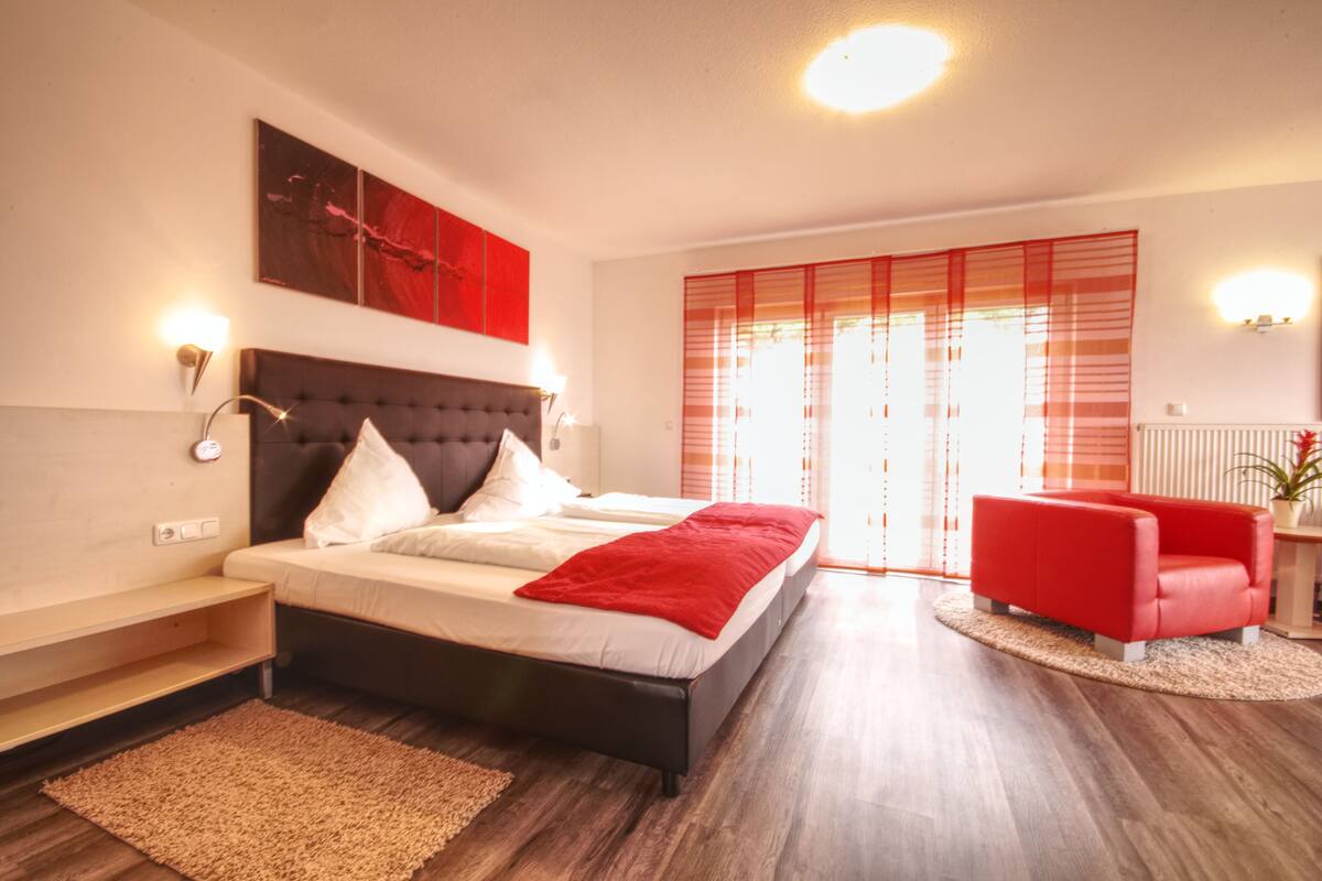 Quarto moderno com uma cama grande, uma cadeira vermelha e vista para o exterior através de uma janela com persianas vermelhas.