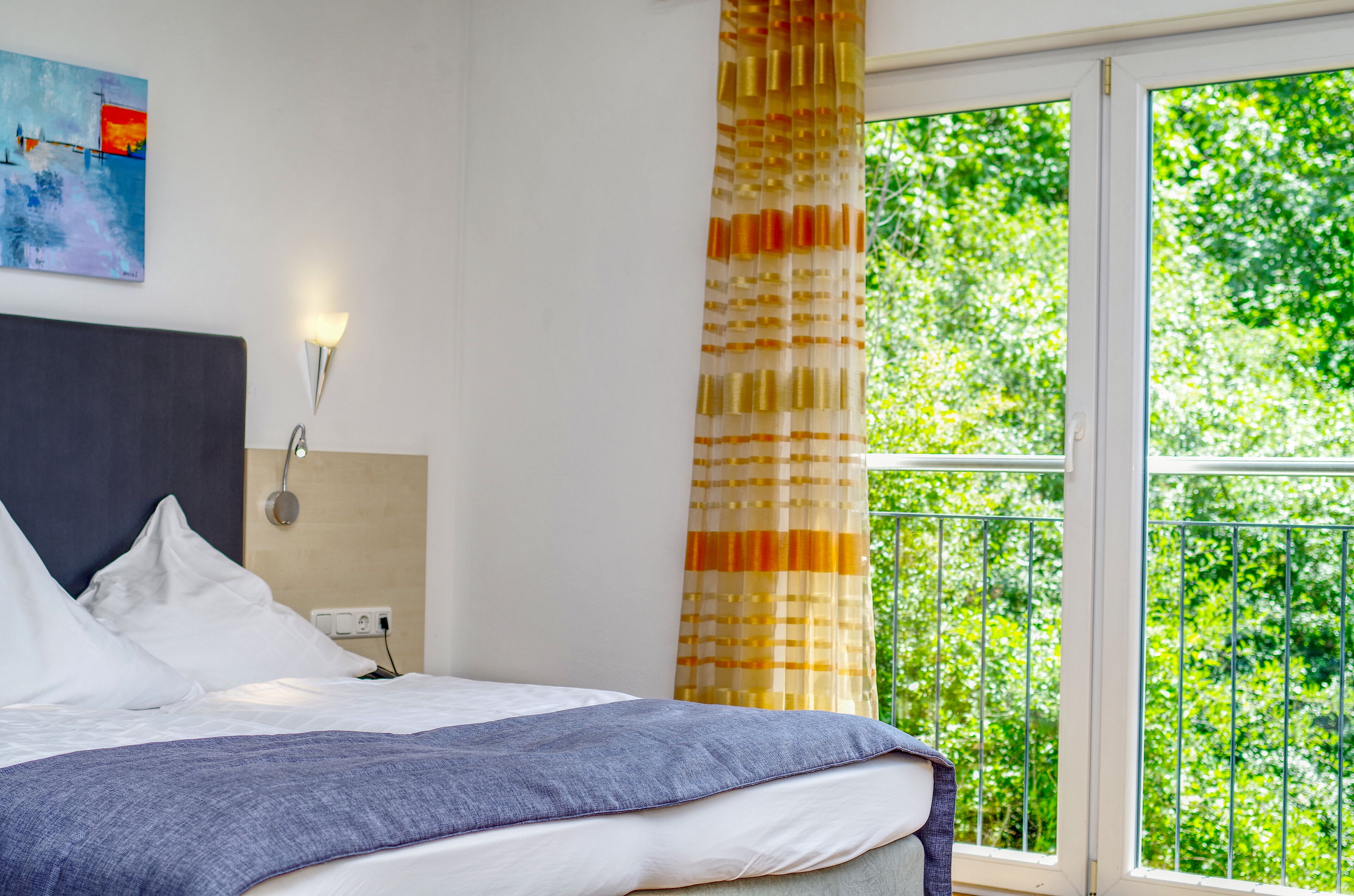 A bedroom with a bed, a nightstand, a wall-mounted lamp, a window with a view of greenery, and a painting on the wall.