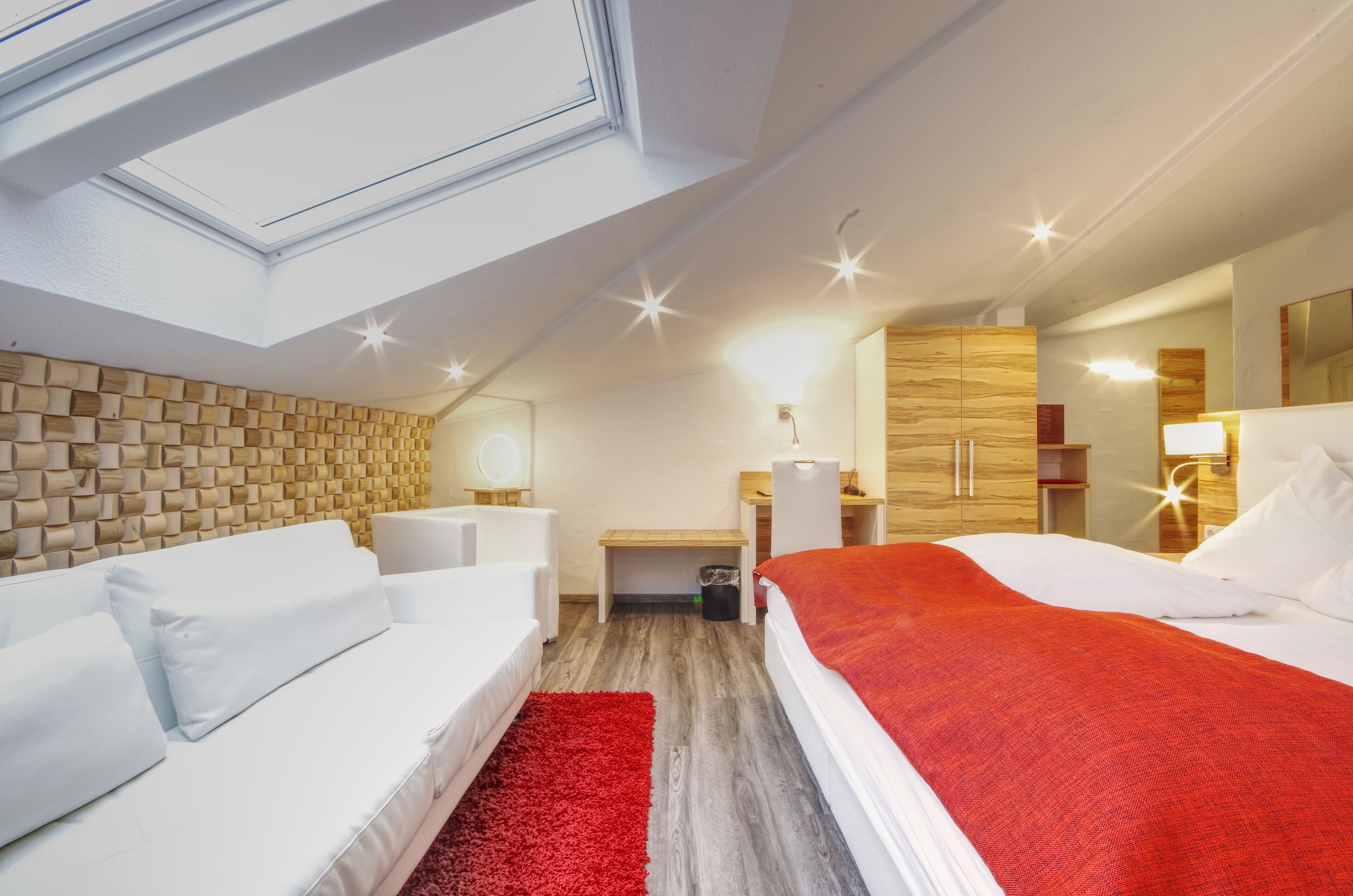 Ein modernes Hotelzimmer mit Dachfenster, einem weißen Sofa, einem roten Teppich, einem Bett mit roter Decke und Holzvertäfelung an den Wänden.