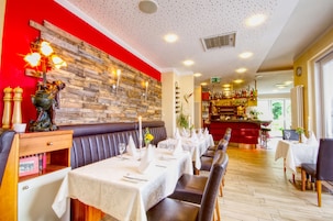 A restaurant with a red accent wall, wooden paneling, and a bar area. Tables are set with white tablecloths and candles.