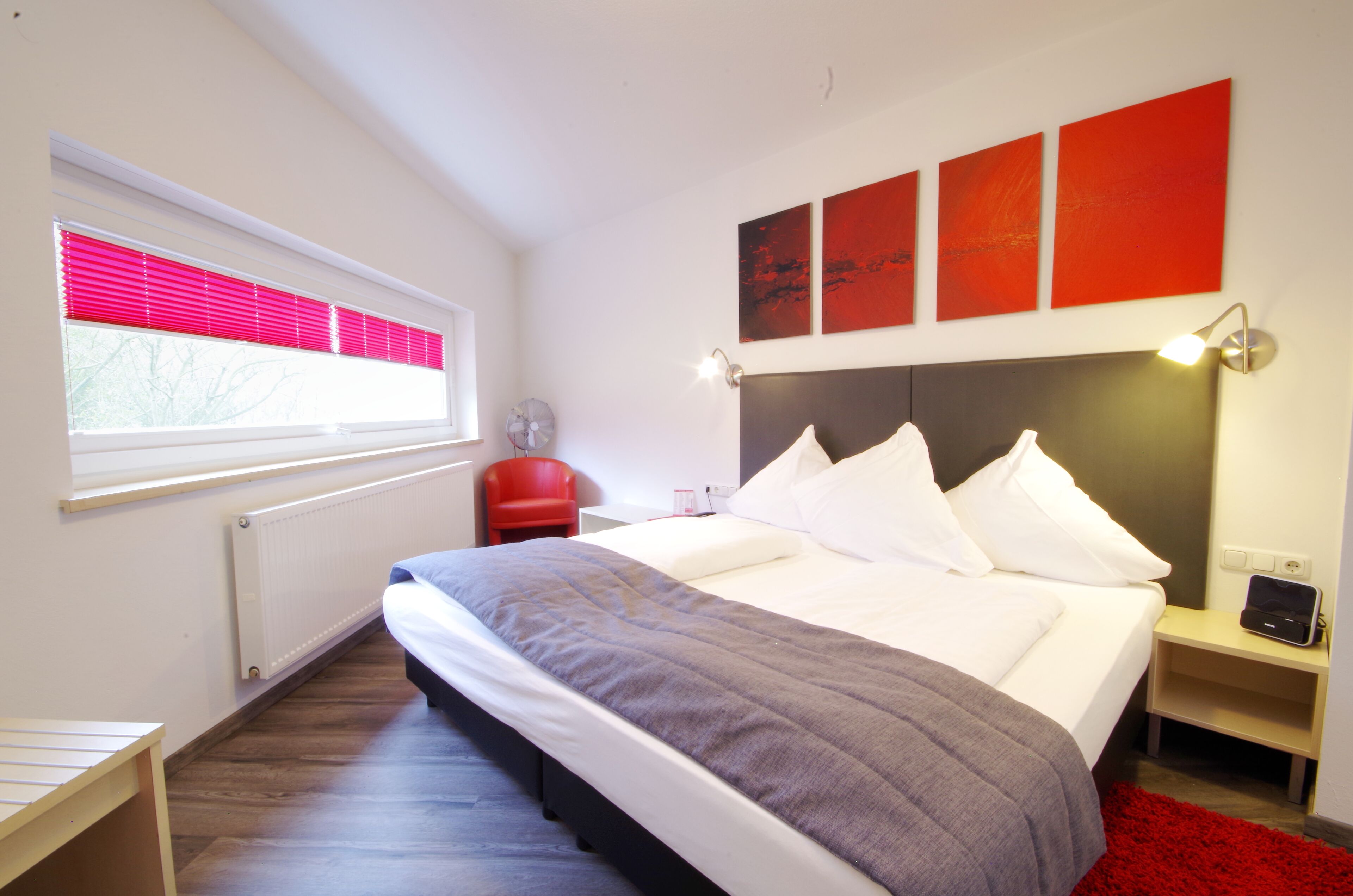 Un dormitorio moderno con una cama grande, una silla roja, una mesita de noche con una lámpara y una ventana con persianas.