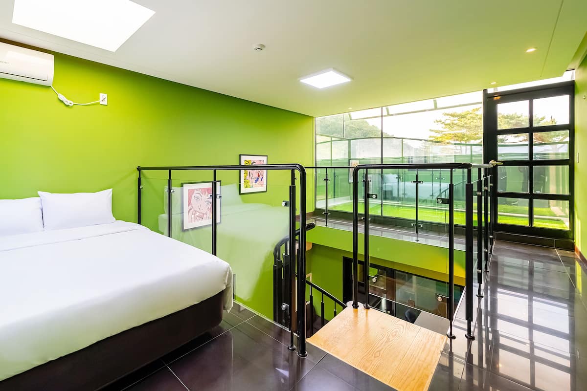 Ein modernes Hotelzimmer mit Bett, grüner Wand, Fenster mit Blick auf einen Tennisplatz und Balkon mit Glasgeländern.