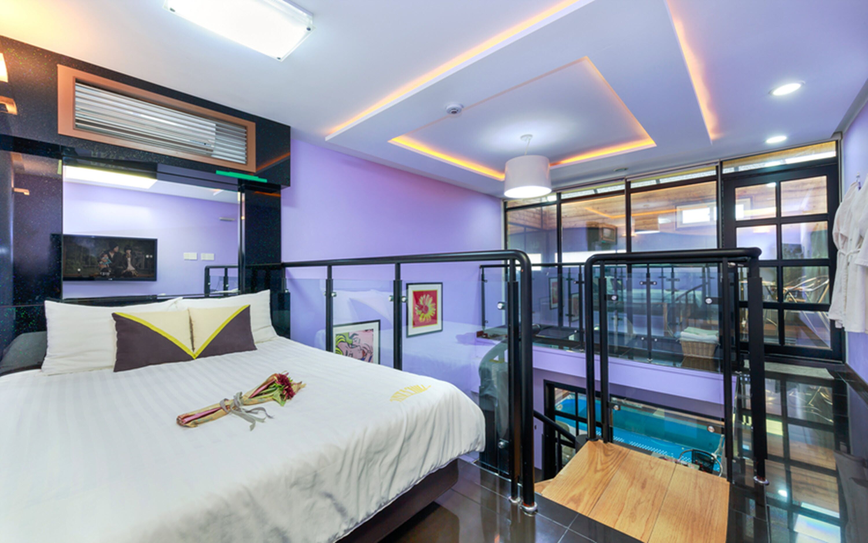 A modern hotel room with a bed, a glass balcony, and a view of a swimming pool.