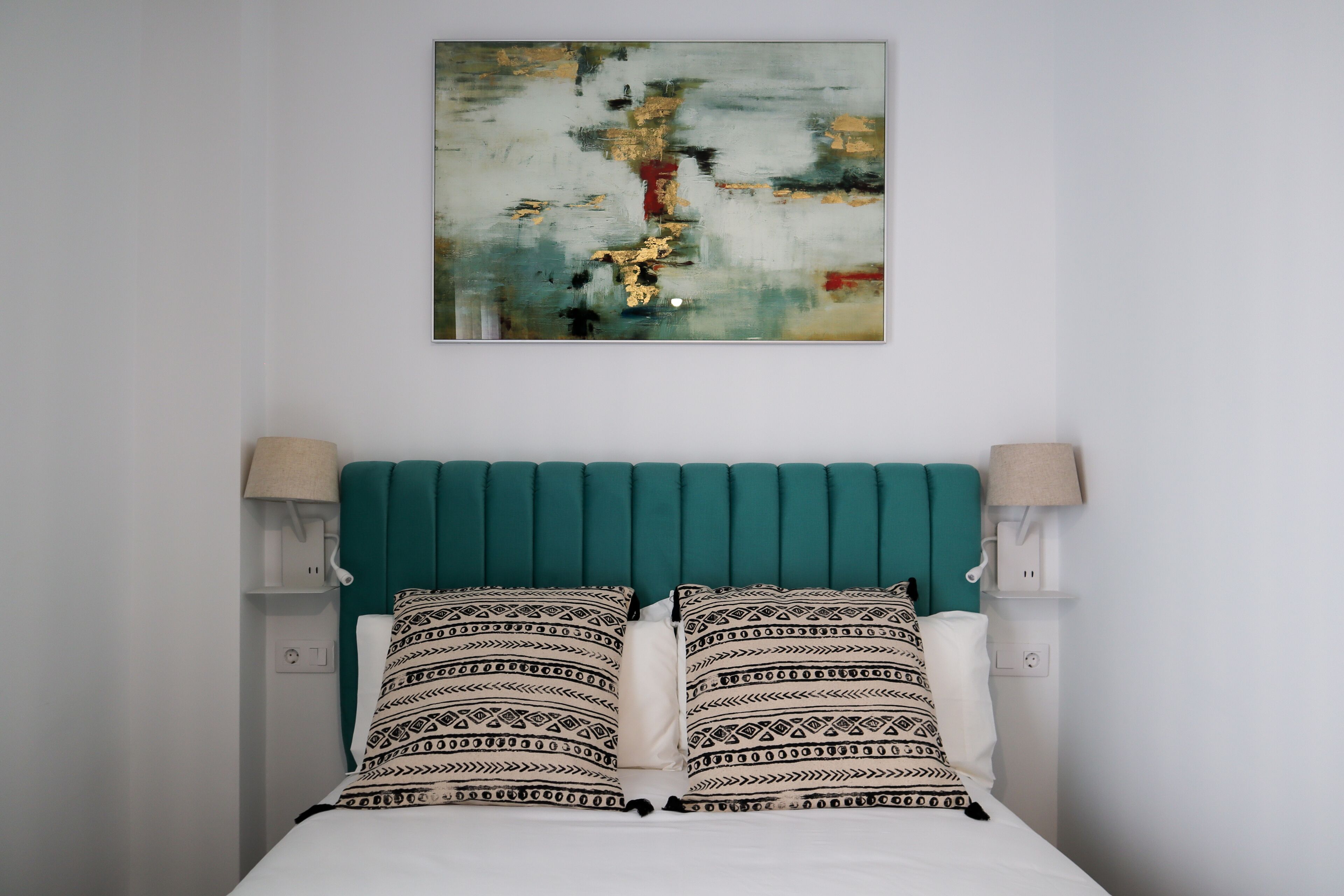 Un lit avec une tête de lit de couleur turquoise, des coussins à motifs et un tableau abstrait accroché au mur.