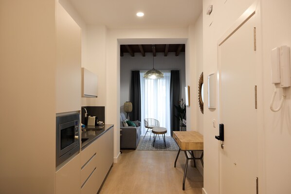 A modern kitchen with a built-in microwave, a dining area with a wooden table and chairs, and a living space with a sofa and a lamp. - Calma Suites (Seville)