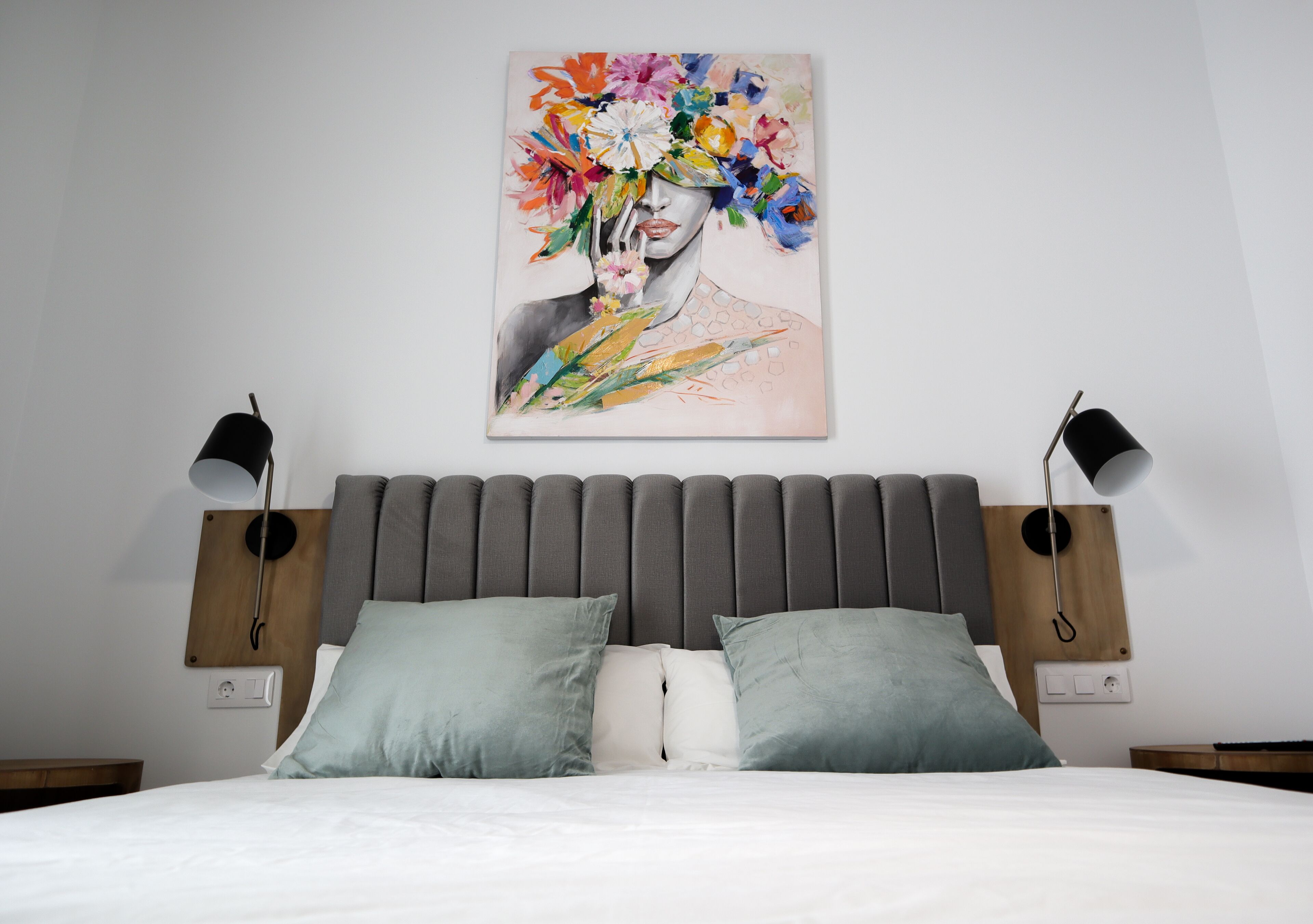 Un dormitorio moderno con un cabecero tapizado gris, una colcha blanca y dos lámparas de noche.