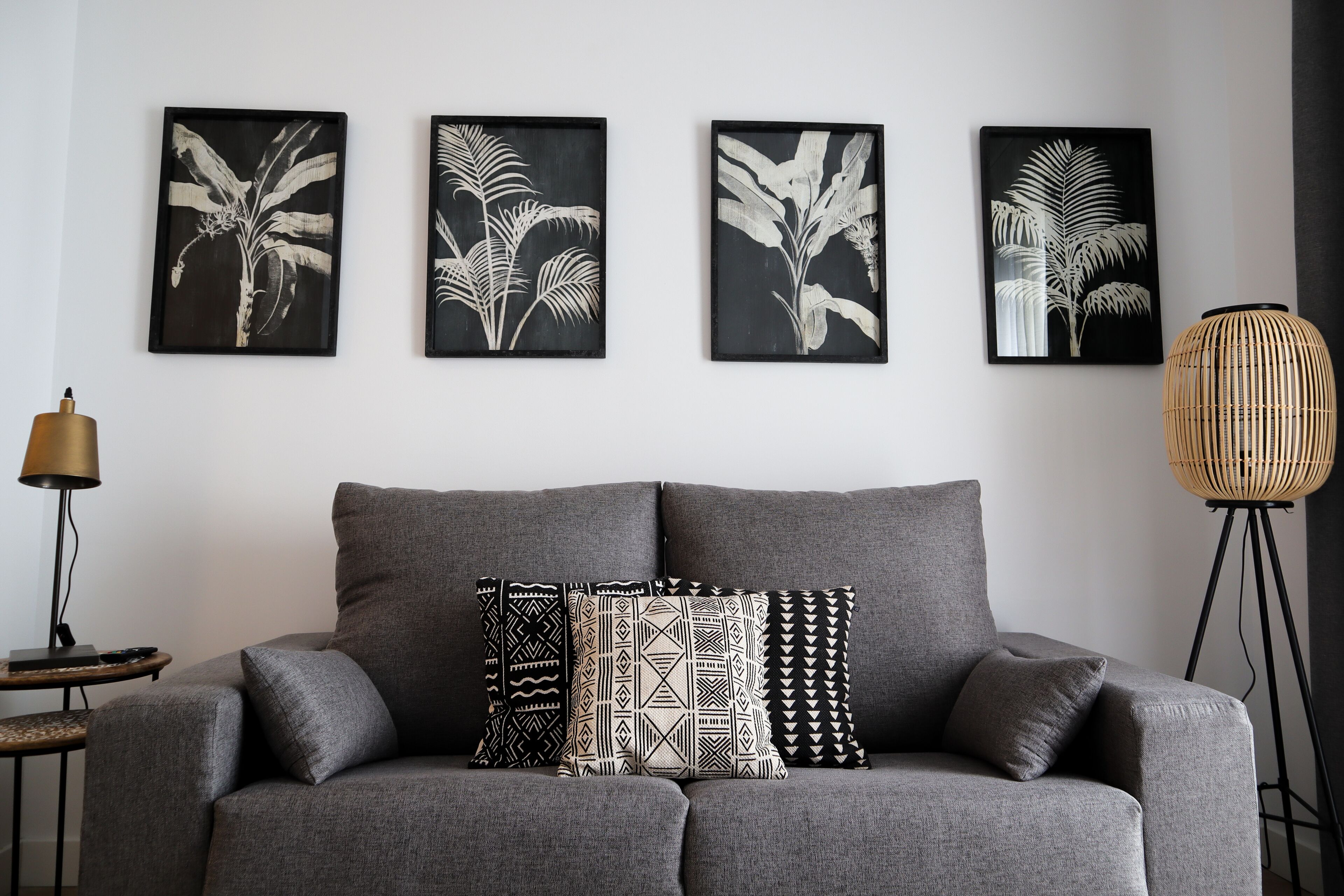 Ein graues Sofa mit gemusterten Kissen, eine Wandkunst mit Blättermotiv in schwarzem Rahmen, eine Stehlampe und eine gewebte Stehlampe.