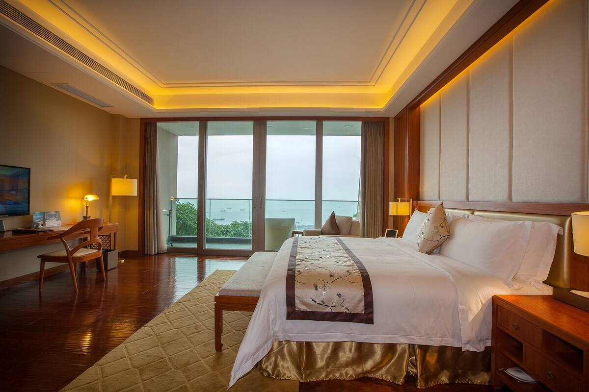 Habitación de hotel con una cama grande, mesitas de noche, un escritorio y una silla. Se ve el mar a través de amplias ventanas.