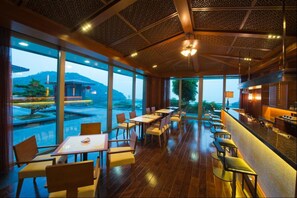A restaurant with wooden flooring, tables, and chairs arranged for dining. Large windows offer views of a mountainous landscape.