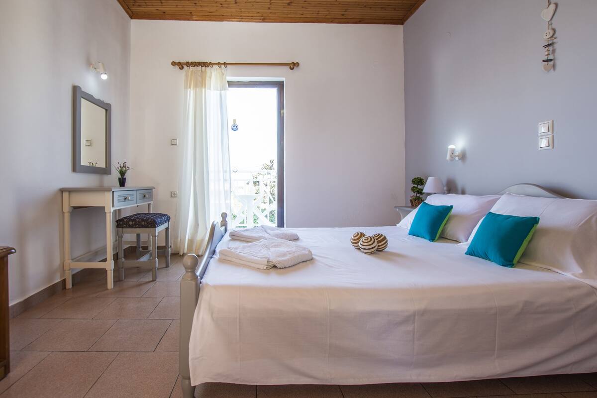 Un dormitorio con cama, mesita de noche, una silla, ventana con cortinas y vistas al exterior.