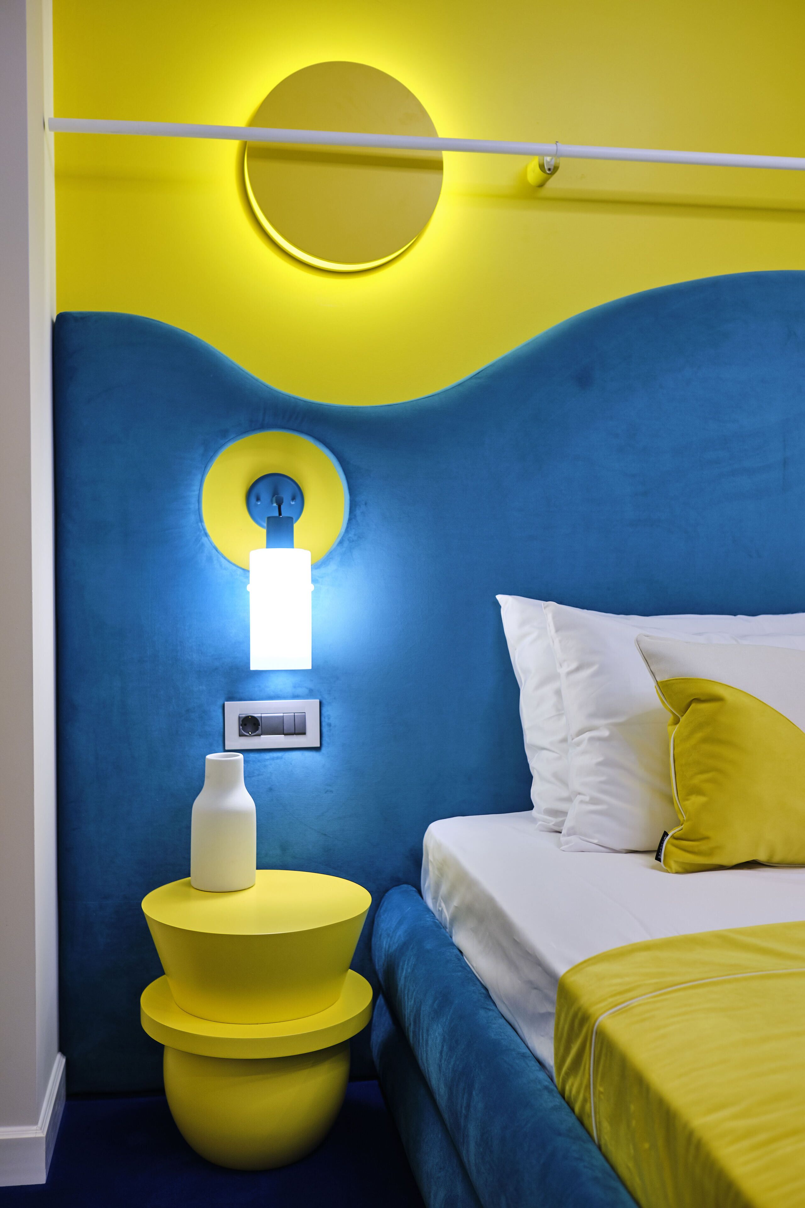 A room with a blue and yellow color scheme, featuring a bed with white and yellow bedding, a bedside table with a white vase, and a wall-mounted light fixture.