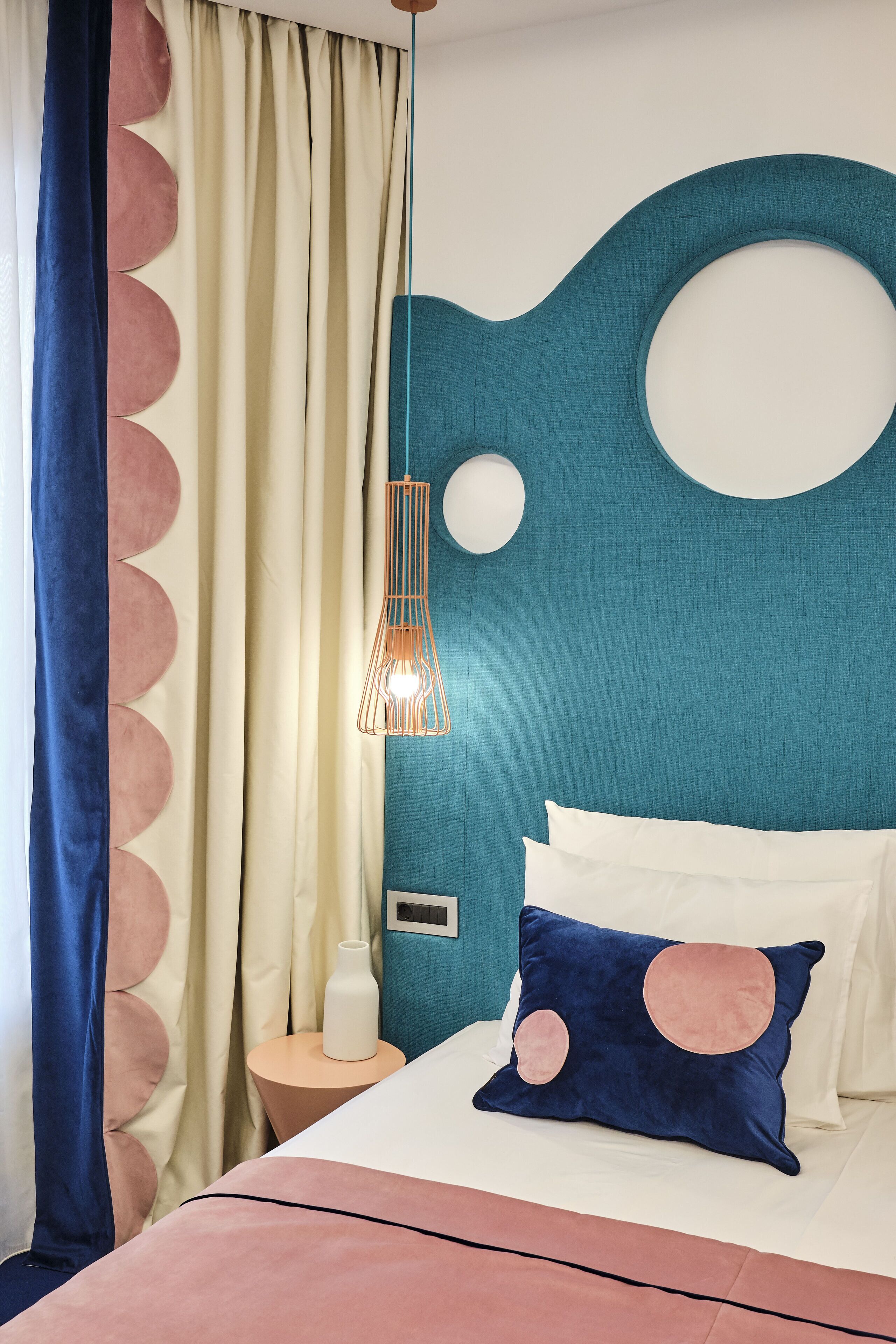 A bedroom with a teal accent wall, a bed with white and pink pillows, a round pendant light, and a side table with a bottle and a vase.