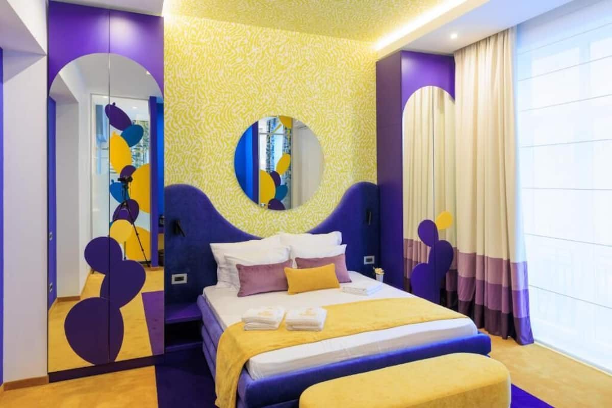 A modern bedroom with a large bed, purple and yellow decor, and a mirror above the bed.